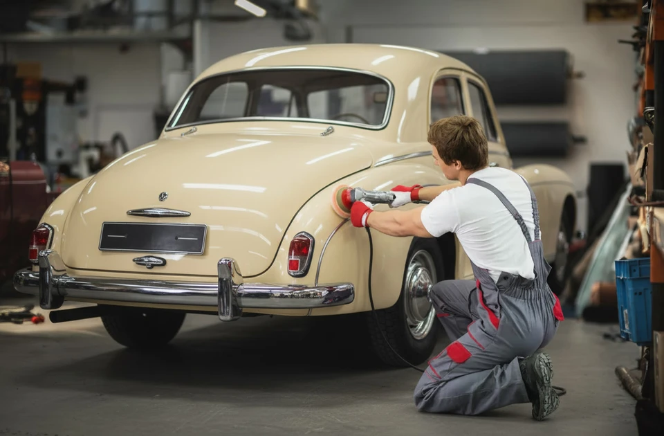Atelier automobile intérieur, technicien en combinaison grise et rouge utilisant une polisseuse pneumatique sur la carrosserie d'un SUV bleu foncé